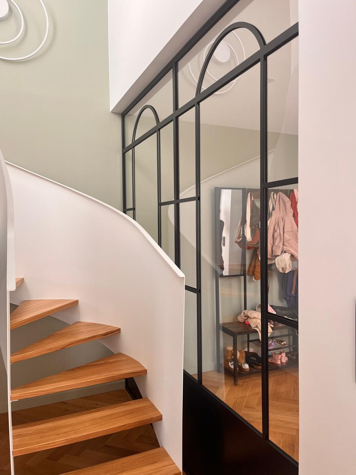 Verrière en acier noir à imposte cintrée, le long d’une volée d’escalier à marches de bois.