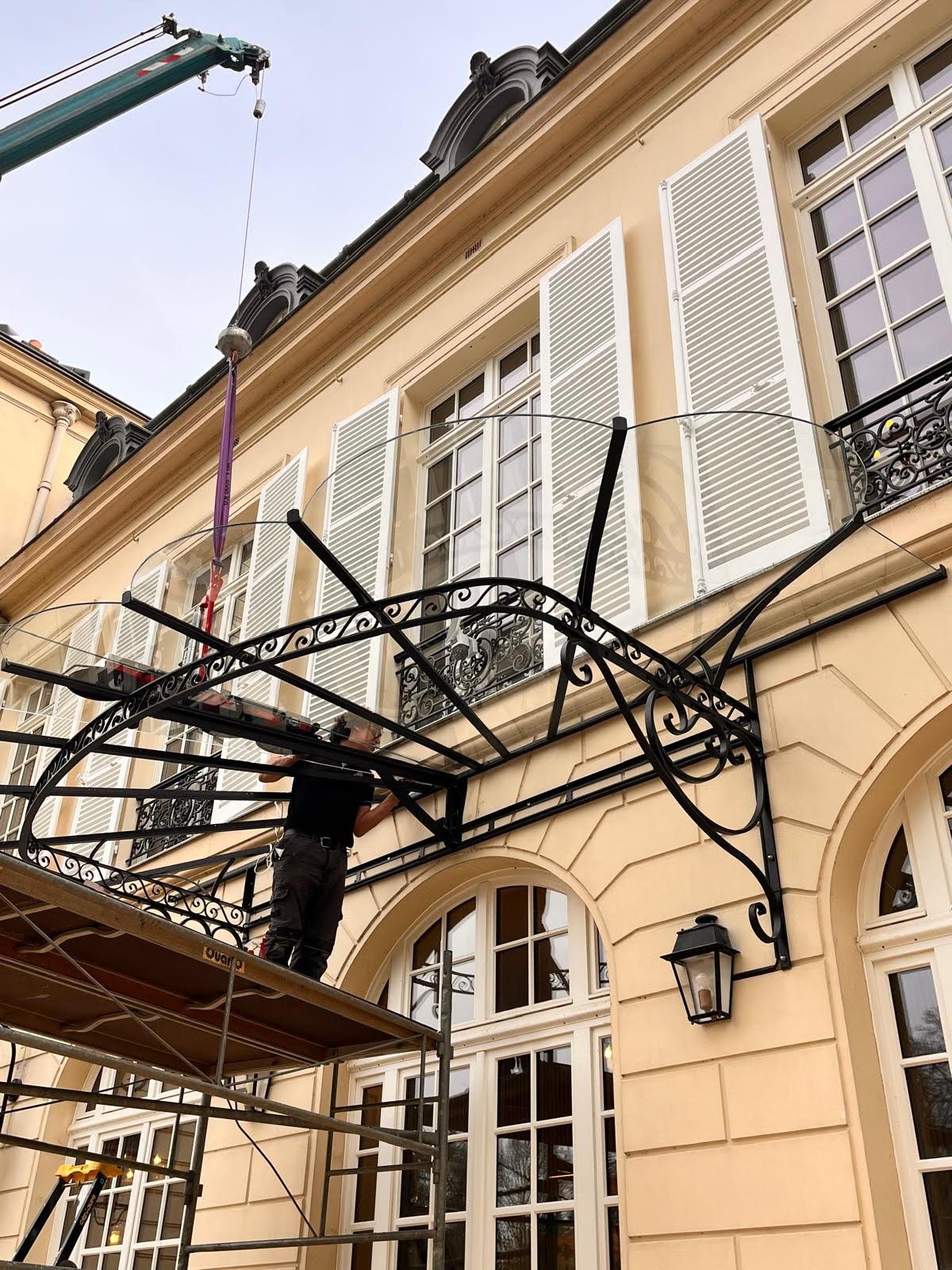 Marquise en fer forgé levée à la grue devant la façade d’un immeuble en pierre de taille.