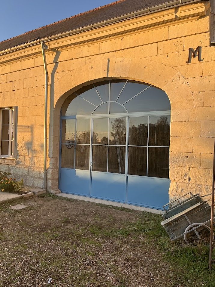 Grande baie cintrée vitrée à meneaux d’acier, ouverte dans une façade de pierre.