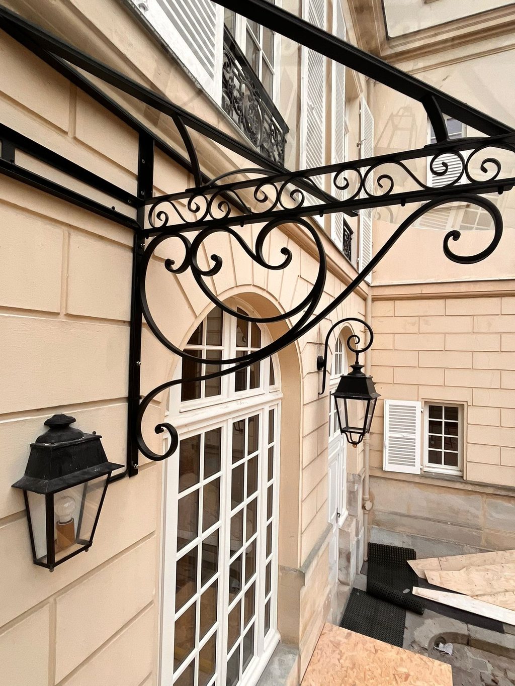 Marquise en fer forgé à volutes au-dessus d’une porte, près d’une lanterne murale.