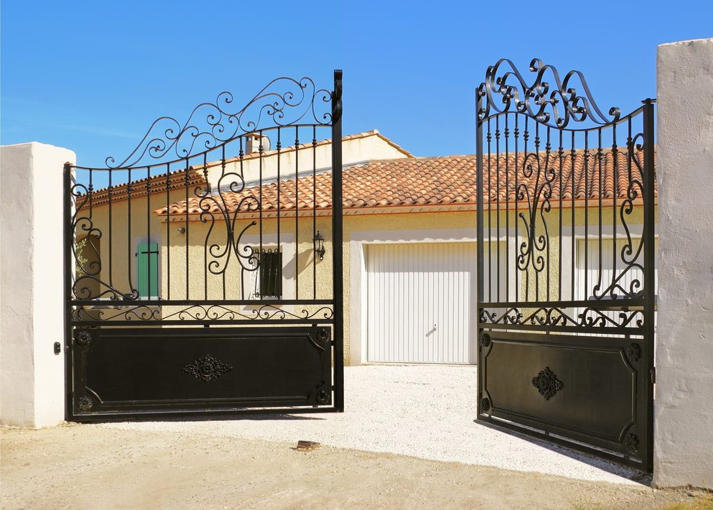 Portail à deux vantaux en fer forgé, motifs volutes, devant une maison.