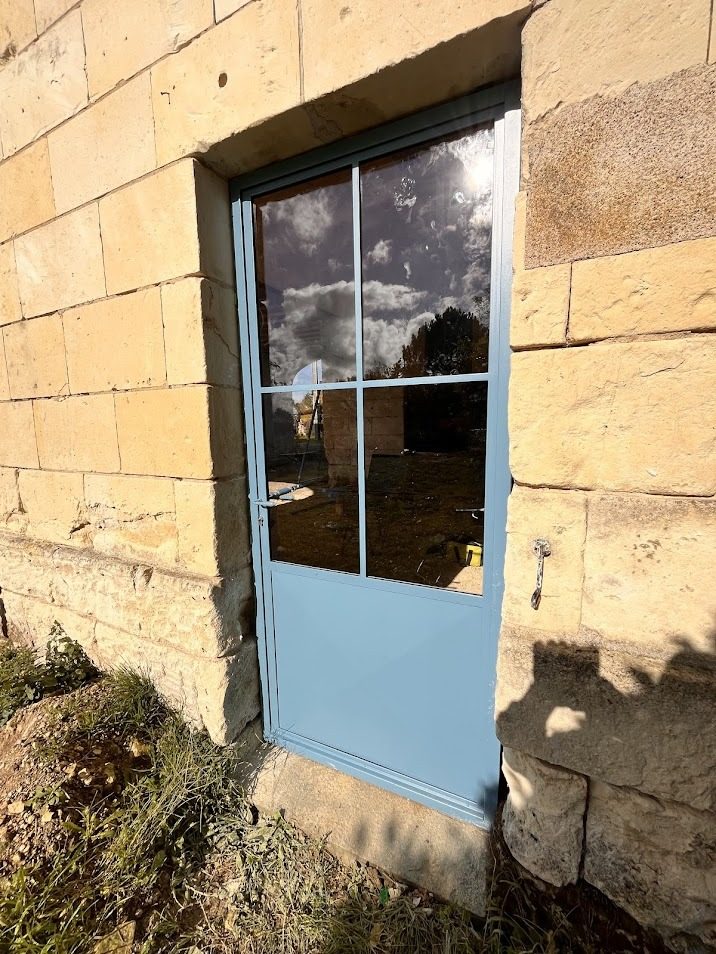 Porte vitrée à cadre d’acier ouverte dans un mur de pierre de taille, en façade.
