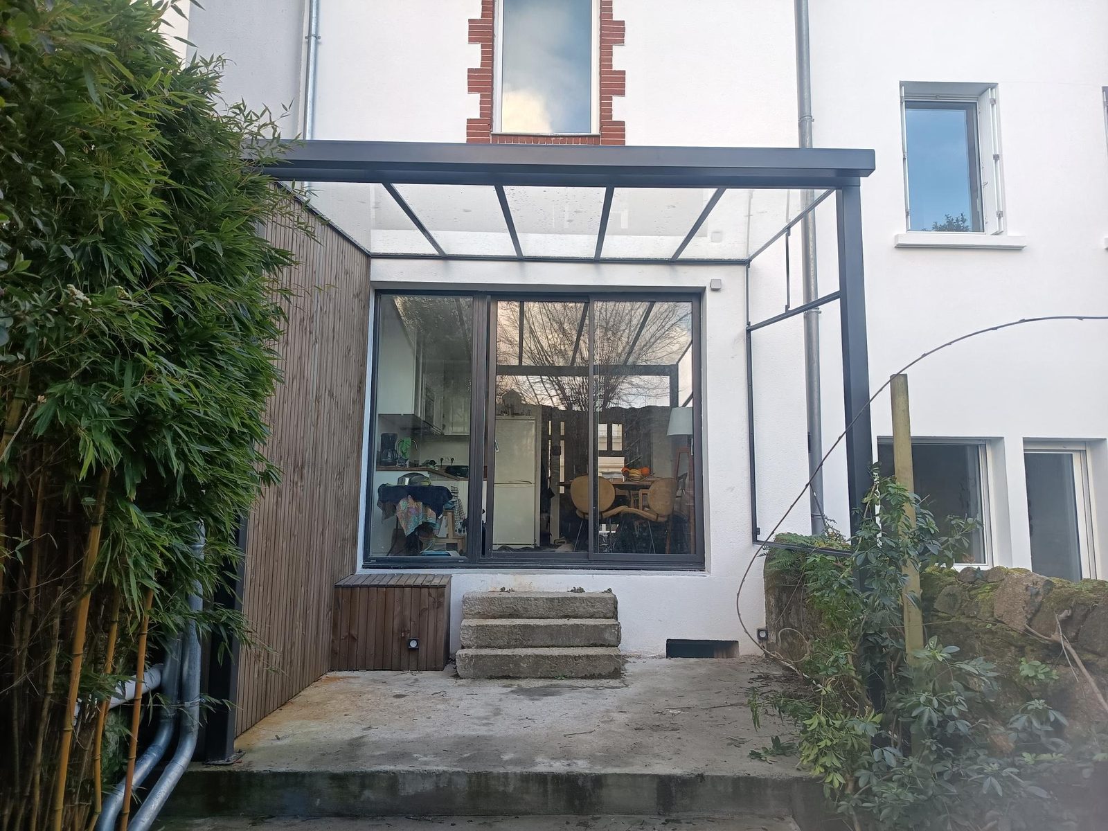 Véranda en acier et verre adossée à une maison, ouverte sur un jardin de bambous.