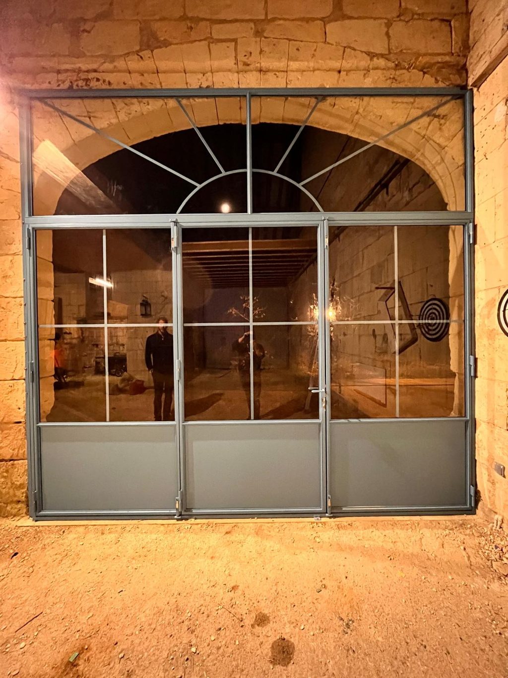 Verrière en acier dans une arche de pierre, éclairée de l’intérieur à la tombée du jour.
