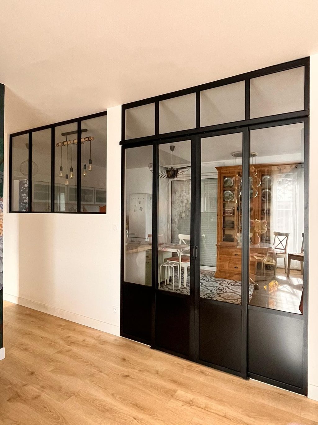 Cloison vitrée en acier noir entre un couloir et une pièce de vie, sur parquet clair.