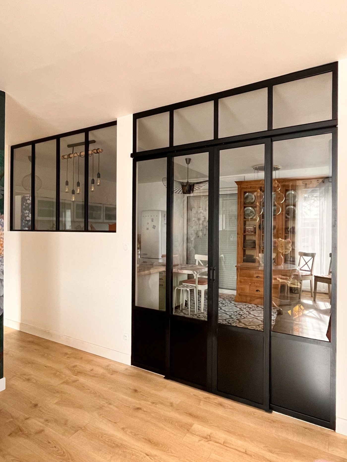 Verrière noire en acier avec portes coulissantes vitrées et impostes, séparant le salon de la cuisine, sur parquet clair.