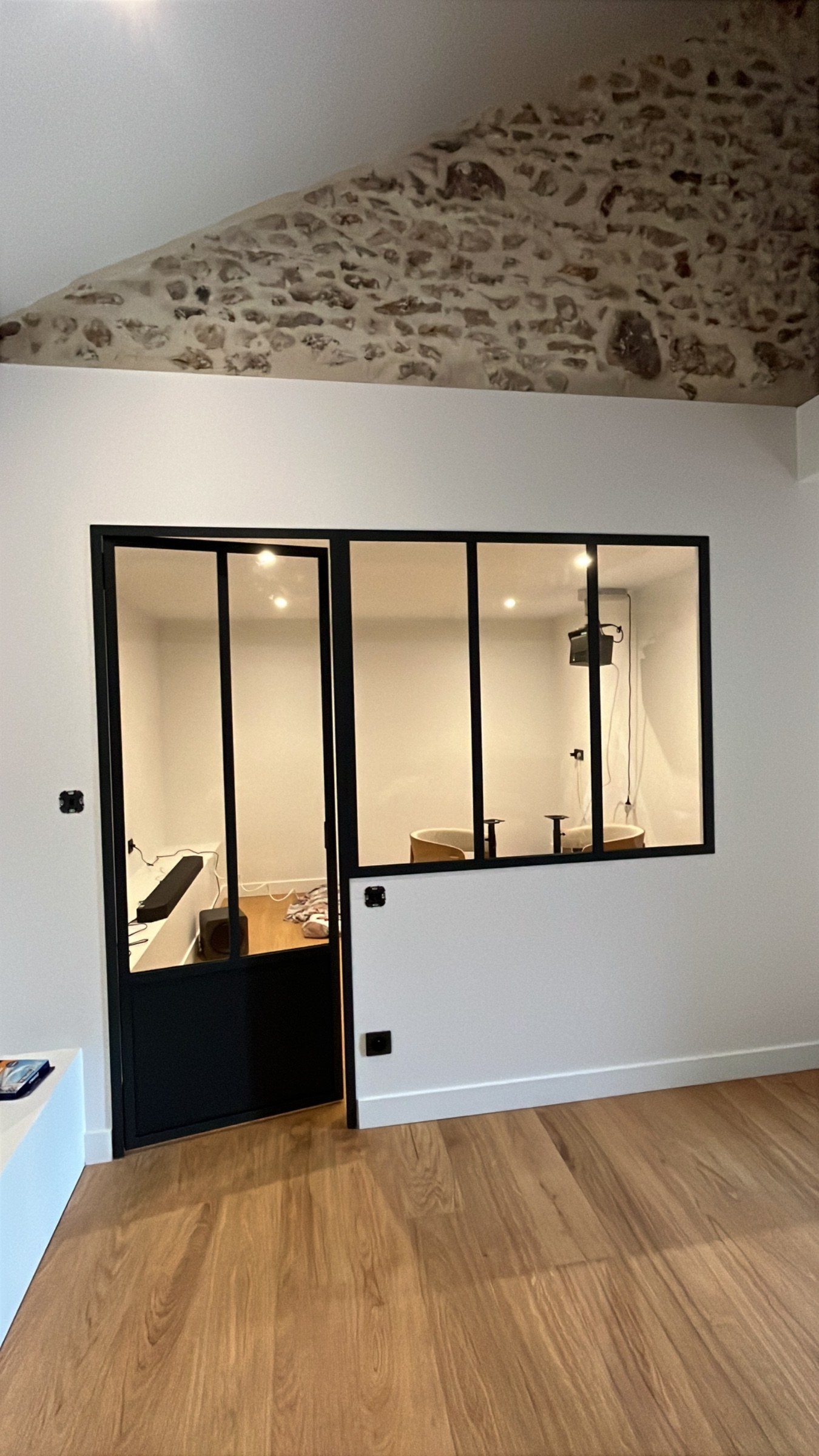 Verrière en acier noir sur mesure avec porte pleine, posée contre un mur blanc sous une charpente en pierre apparente.